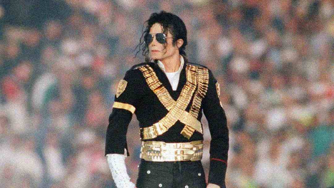 A performer in a black and gold military-style jacket, black pants, sunglasses, and a single white glove stands on stage with a blurred crowd in the background.