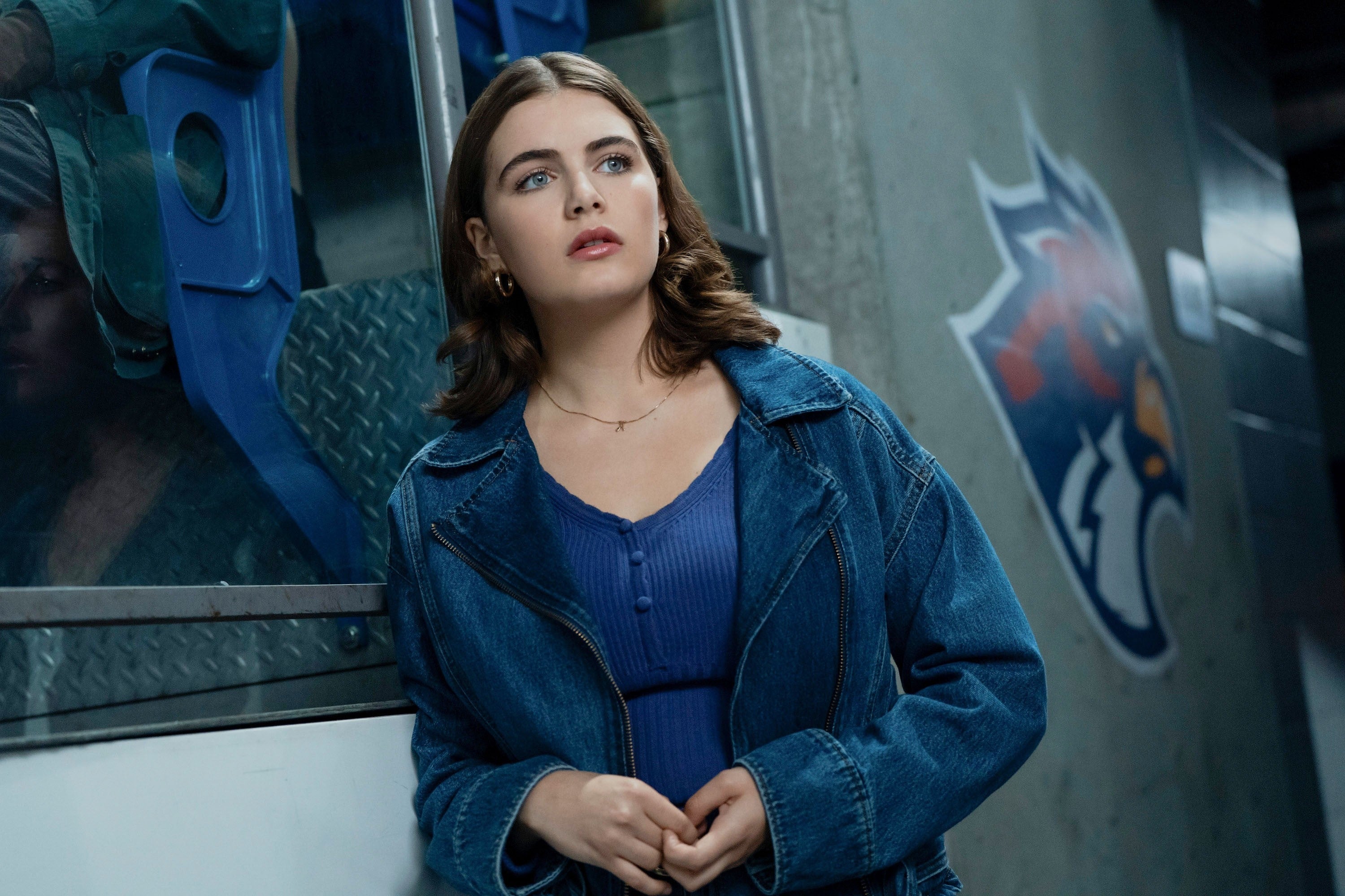 A woman in casual denim attire, with shoulder-length hair, stands pensively by an ice hockey rink, with a team logo visible in the background