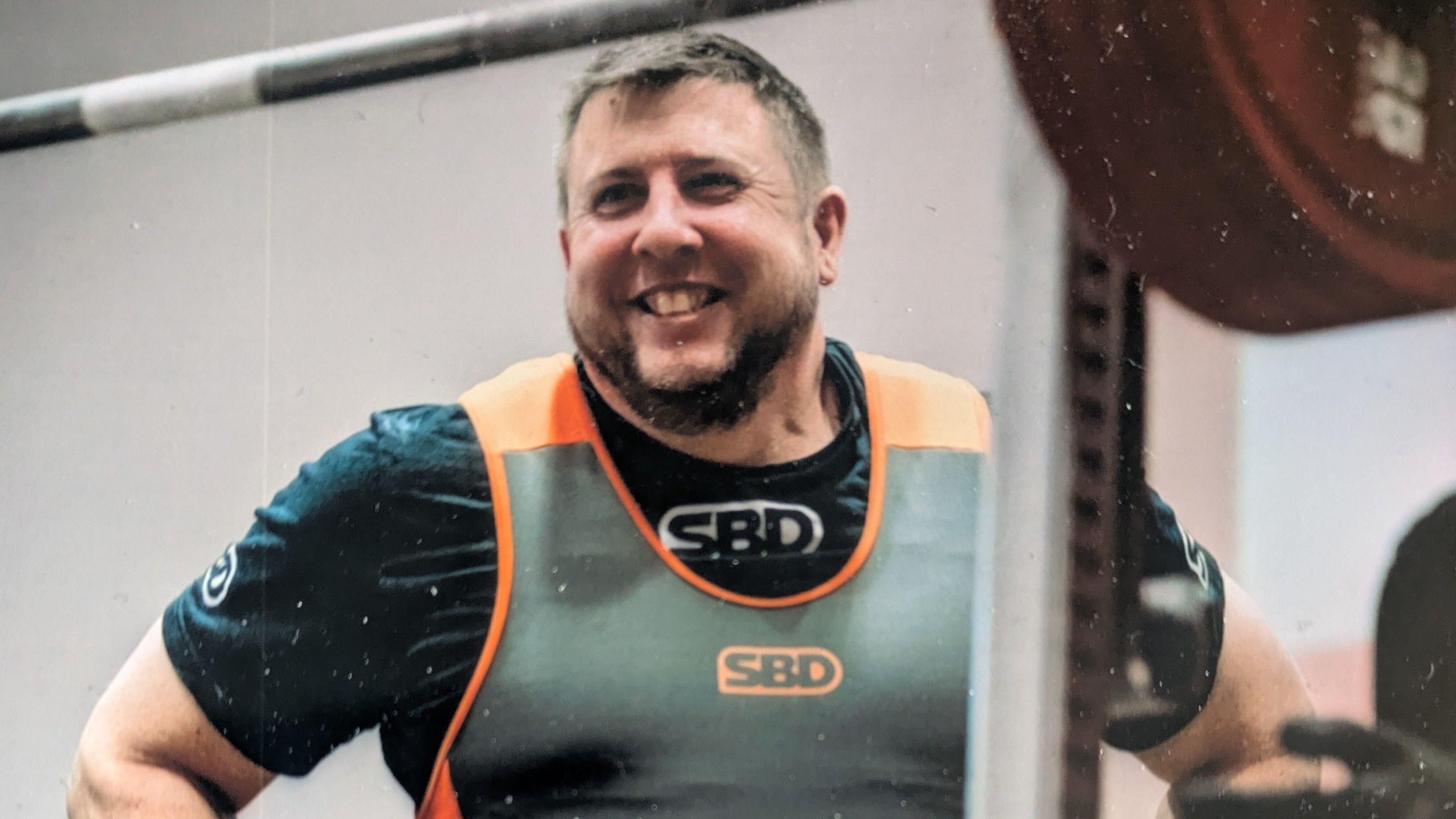 A head and shoulders picture of Chris Pryor wearing a black t-shirt and orange and grey athletic vest. He is standing with his hands on his hips and smiling. There is a large barbell out of focus in the foreground.