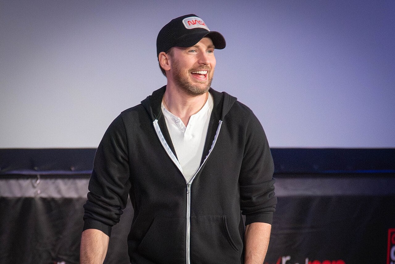 A man wearing a black zip-up hoodie, white shirt, and black baseball cap smiles while standing on stage against a blurred background.