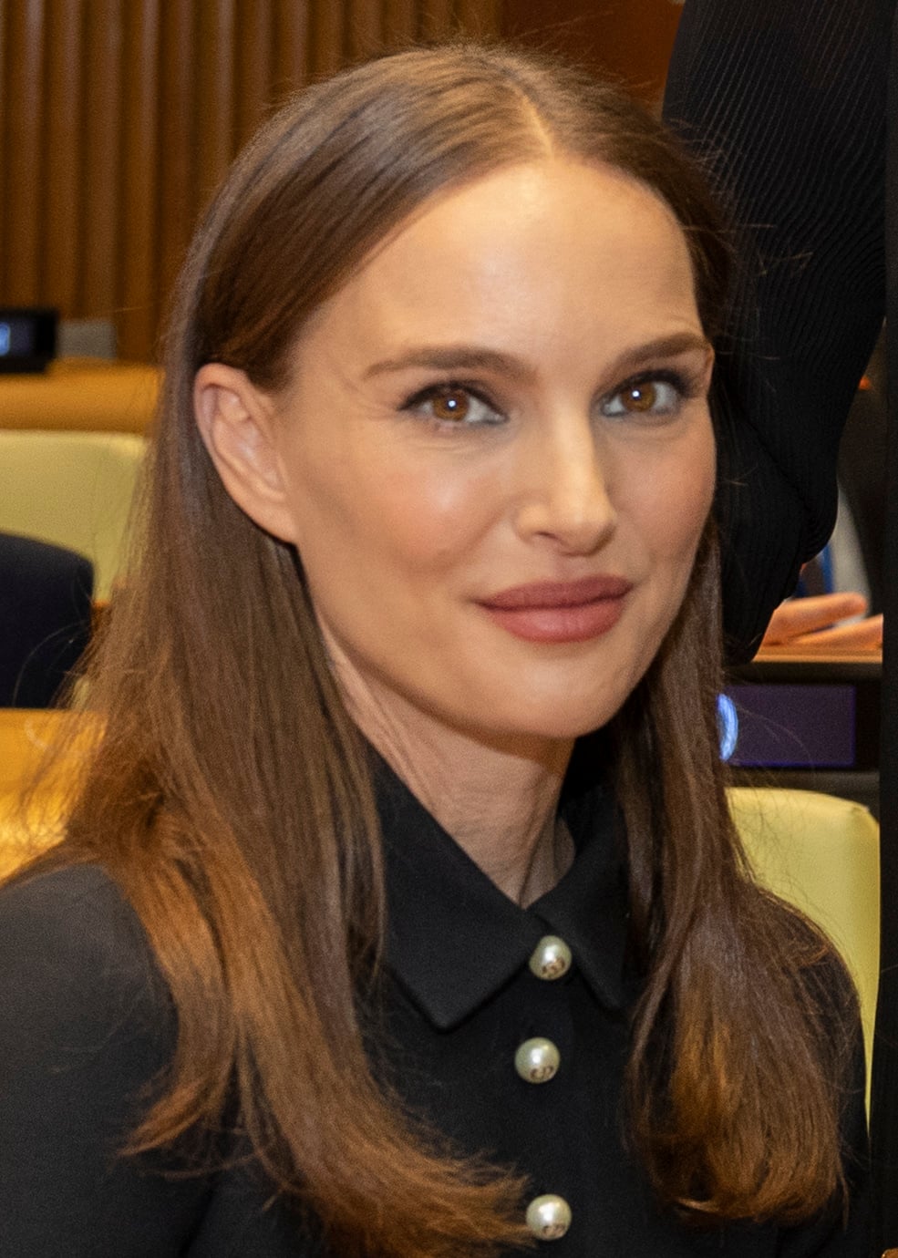 A woman with long straight brown hair, wearing a black collared top with pearl buttons, sits indoors and smiles slightly at the camera. The background shows beige chairs and wood paneling.