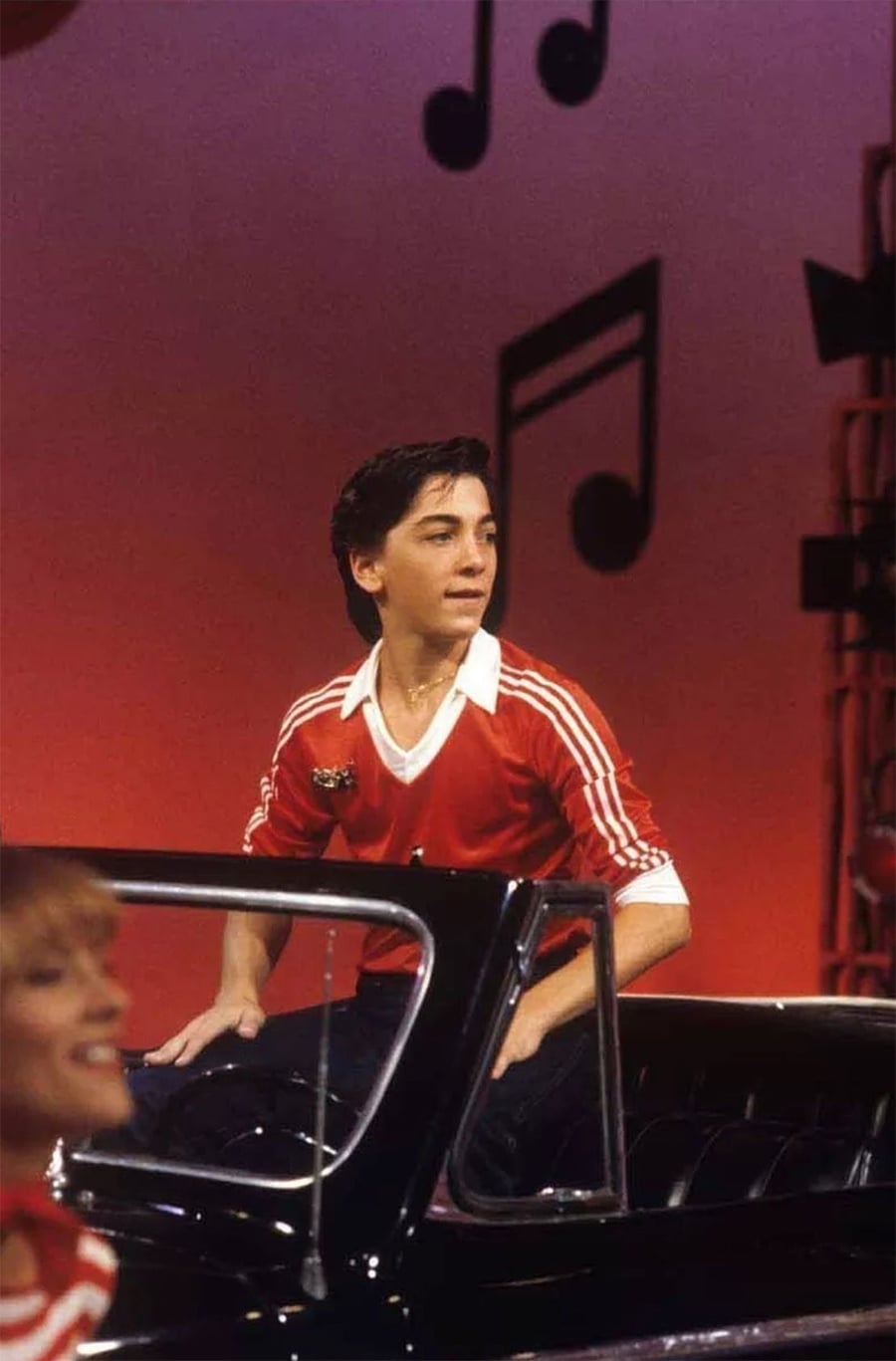 Um jovem com uma camisa listrada vermelha e branca está sentado na traseira de um carro conversível preto em um palco com fundo vermelho e grandes notas musicais pretas penduradas como decoração.