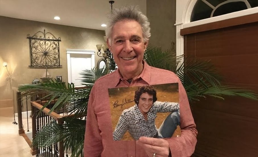 Um homem mais velho com uma camisa vermelha está dentro de casa, sorrindo e segurando uma foto de seu eu mais jovem sentado ao ar livre na grama, vestindo uma camisa xadrez e sorrindo. A sala tem plantas, uma escada e iluminação aconchegante.