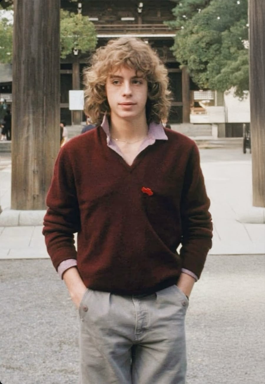 Um jovem de cabelos loiros cacheados, vestindo um suéter marrom sobre uma camisa de colarinho e calça cinza claro, está ao ar livre com as mãos nos bolsos. Grandes pilares de madeira e árvores são visíveis ao fundo.