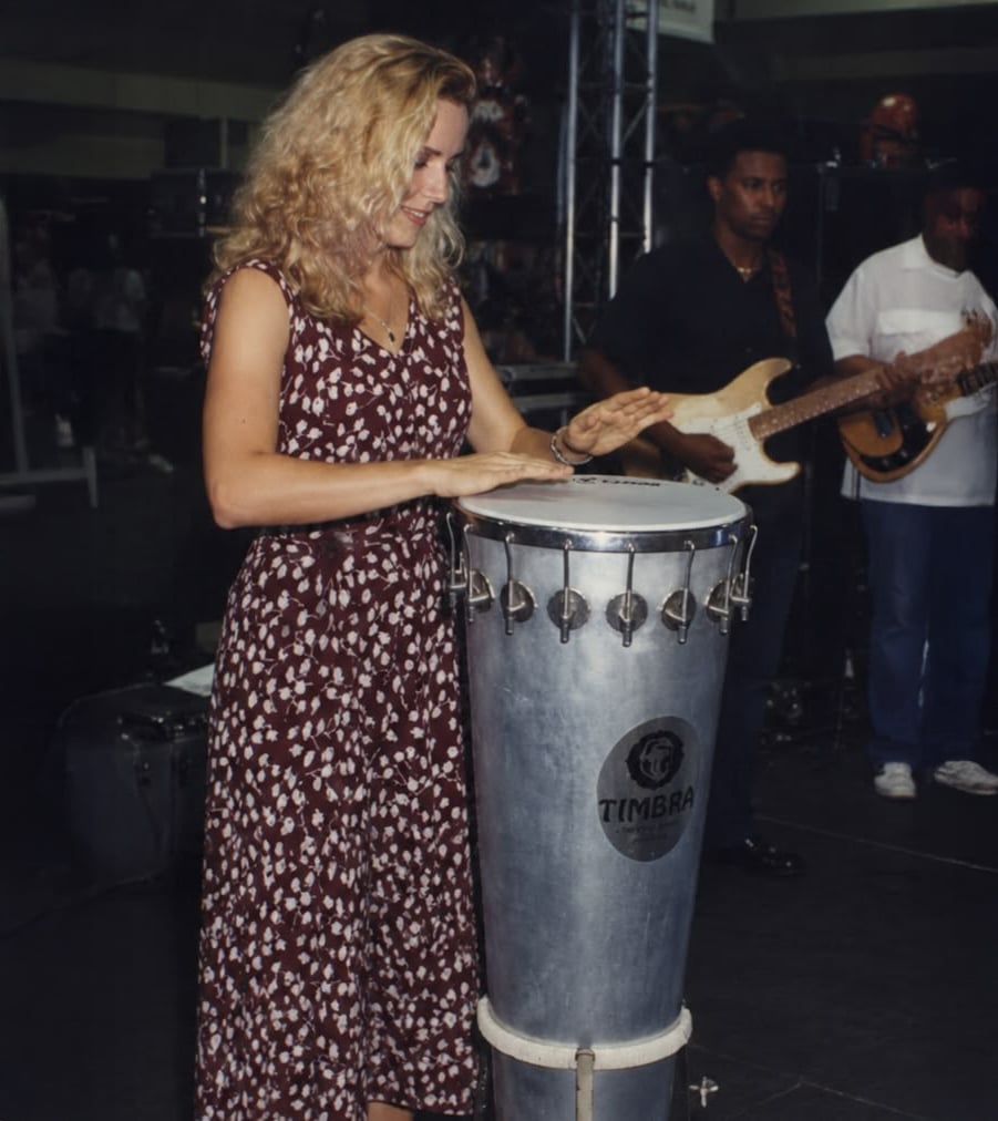 Uma mulher com um vestido estampado toca uma bateria alta no palco, com dois músicos tocando guitarra e baixo ao fundo.