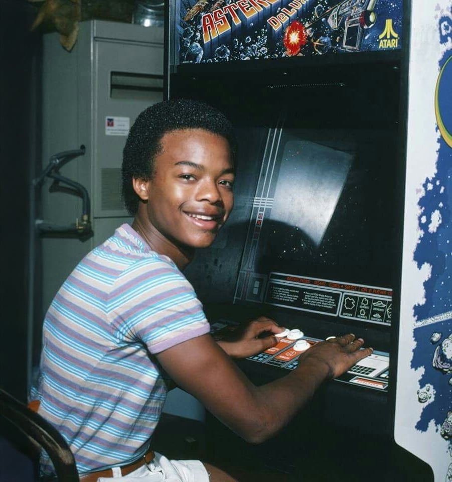 Um adolescente sorridente vestindo uma camisa listrada joga um jogo de arcade Asteroids Deluxe, usando os controles com as duas mãos.