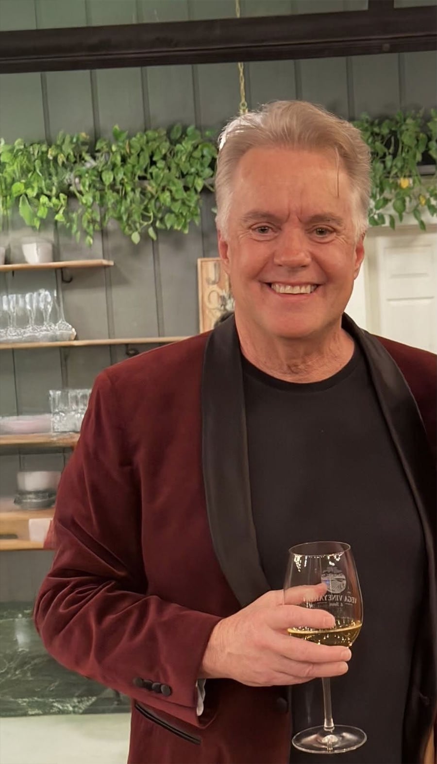Um homem sorridente, com uma jaqueta de veludo marrom e camisa preta, segura uma taça de vinho branco. Atrás dele estão prateleiras com copos e plantas em uma parede verde de madeira.