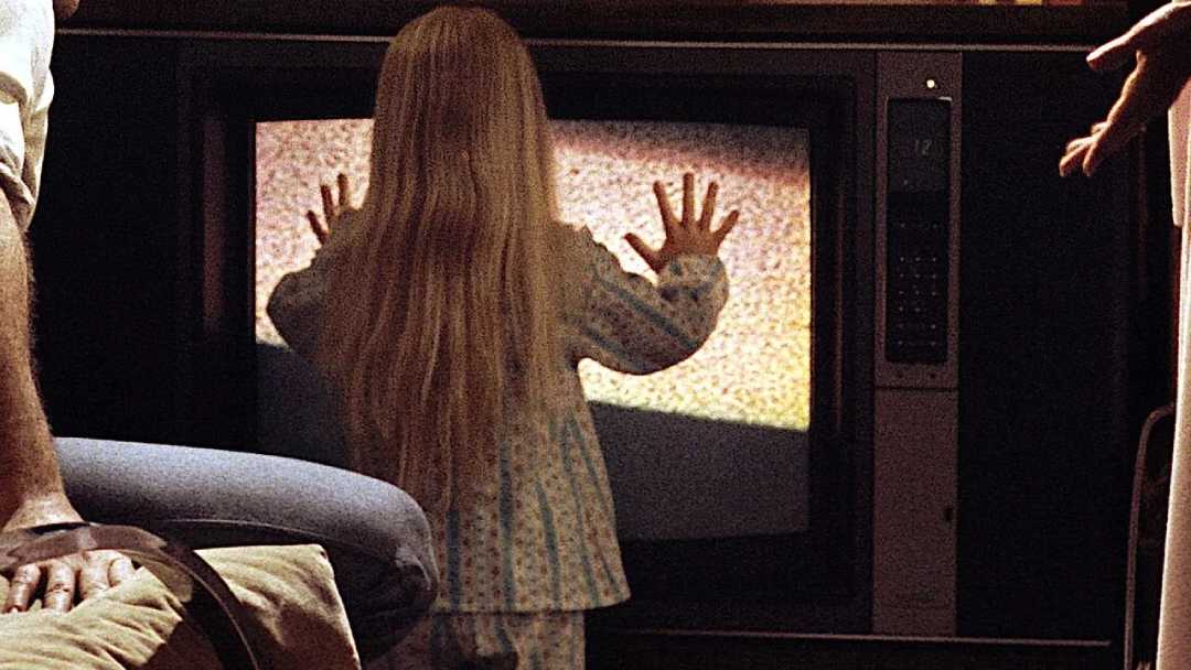 A young girl with long blonde hair touches the static-filled screen of an old television, standing closely in a dimly lit room, while adults are partially visible nearby.
