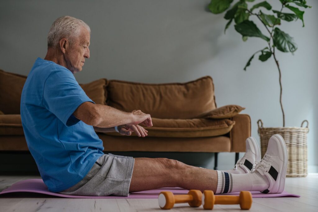 An elderly man wearing a blue shirt is exercising on a yoga mat with dumbbells indoors.