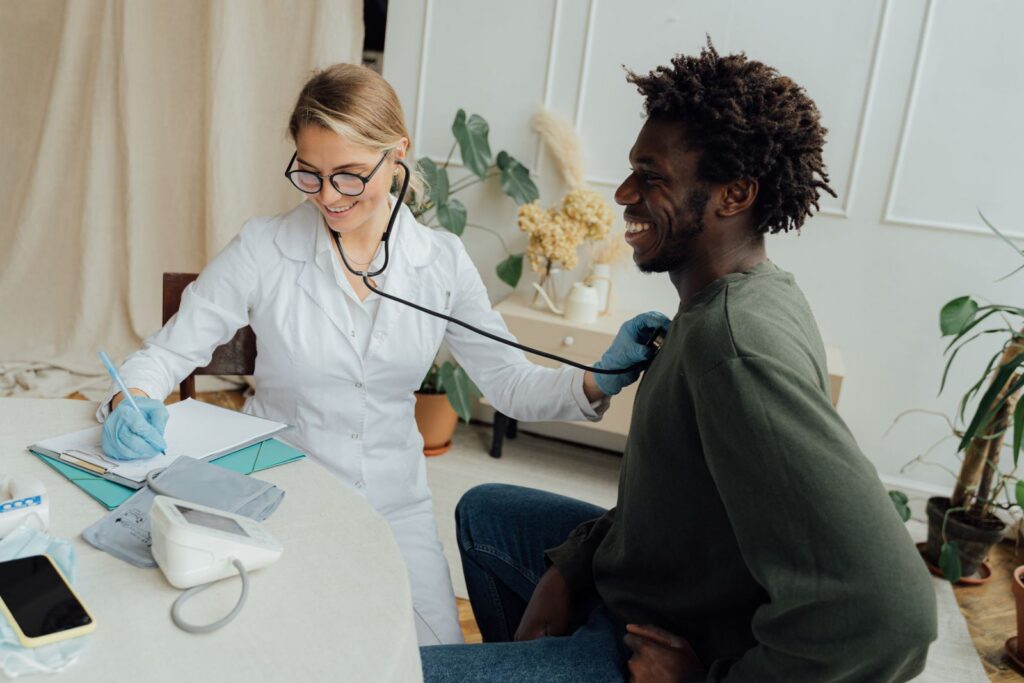 Caucasian female doctor checks the heart rate of a black male patient with a stethoscope in a clinic.