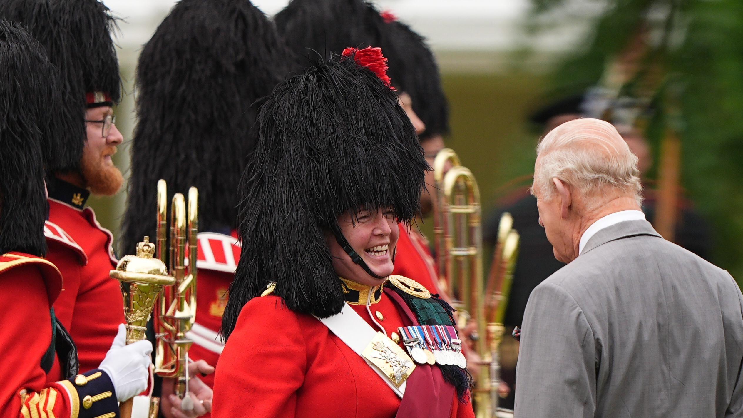 King Charles III (right) during the Ceremony of the Keys in the gardens of the Palace of Holyroodhouse in Edinburgh. He has his back to the camera. Facing the camera are members of the band of the Royal Regiment of Scotland. They are wearing red and gold jackets, a black feather bonnet and are carrying gold instruments.