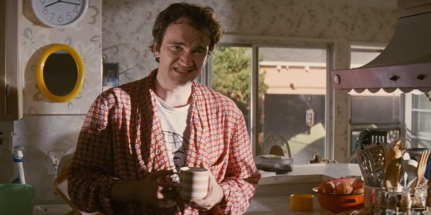 A man in a patterned robe stands in a bright kitchen, holding a striped mug and smiling. Sunlight streams through windows behind him onto a counter with fruit and kitchen utensils.