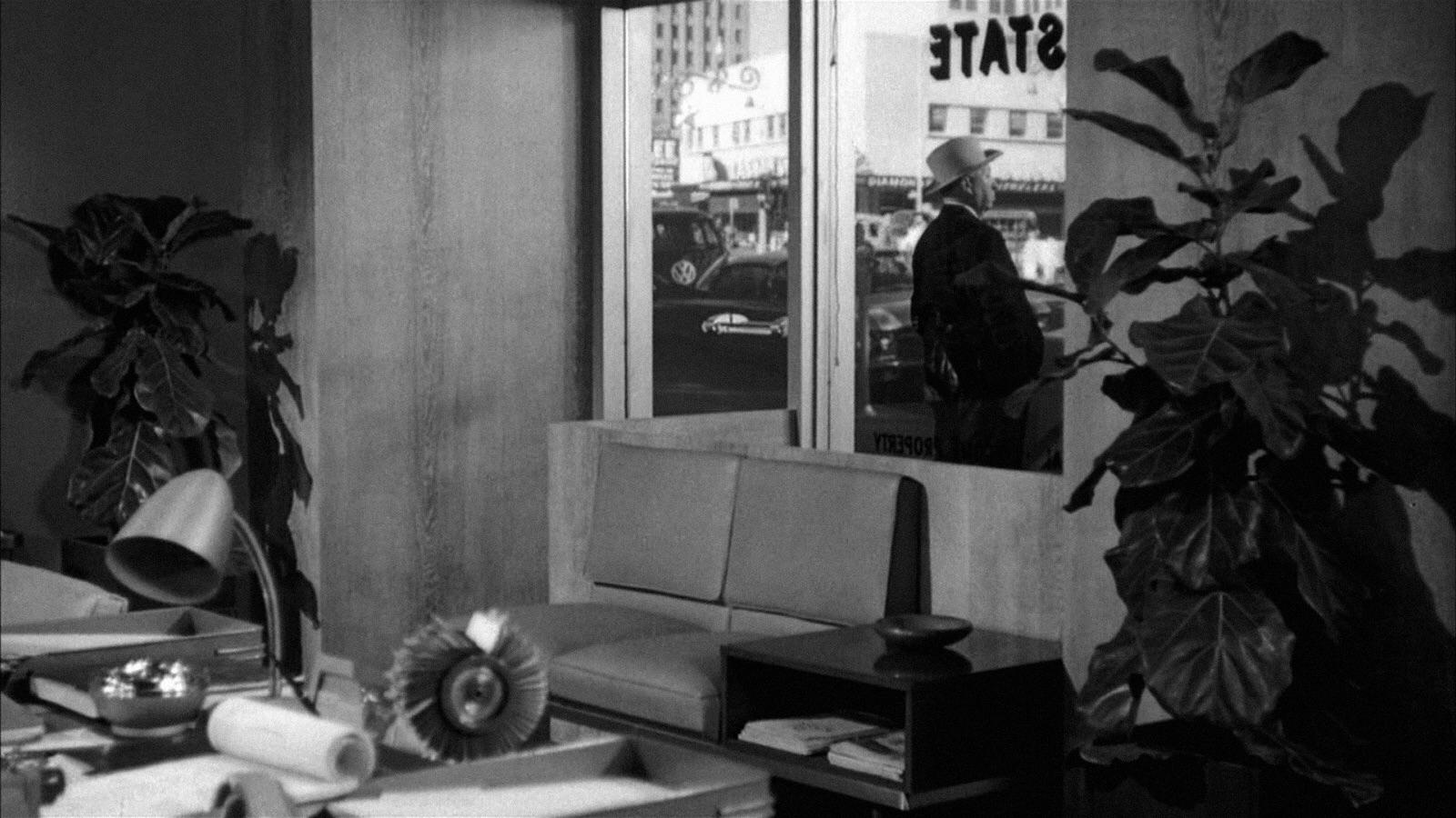 A black-and-white image of a modern office with large plants, a desk with various objects, a couch, and a window. Outside the window, a person in a hat stands on the sidewalk with buildings and cars visible across the street.