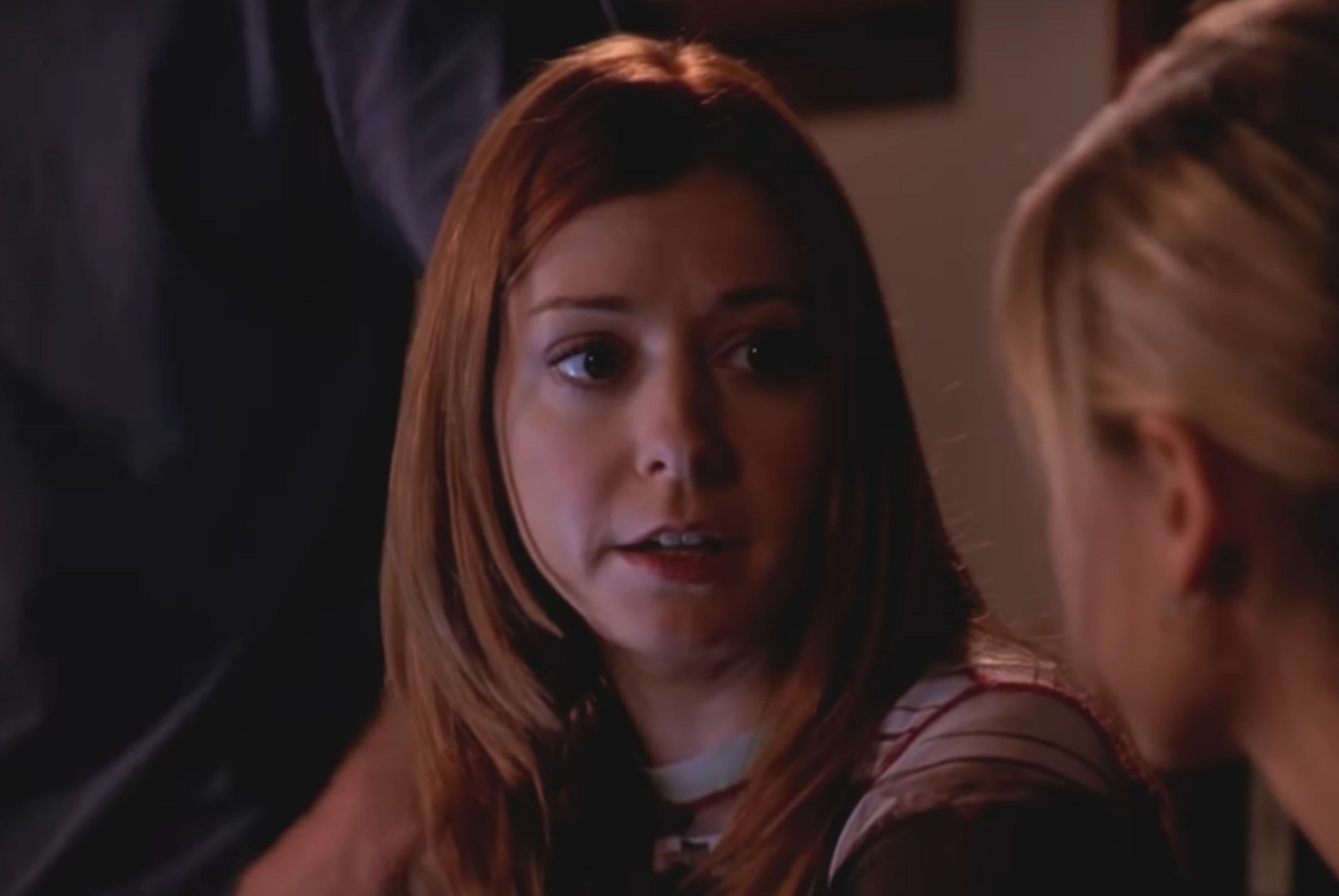 A woman with long hair talks to another person in a dimly lit setting, possibly a scene from a TV show