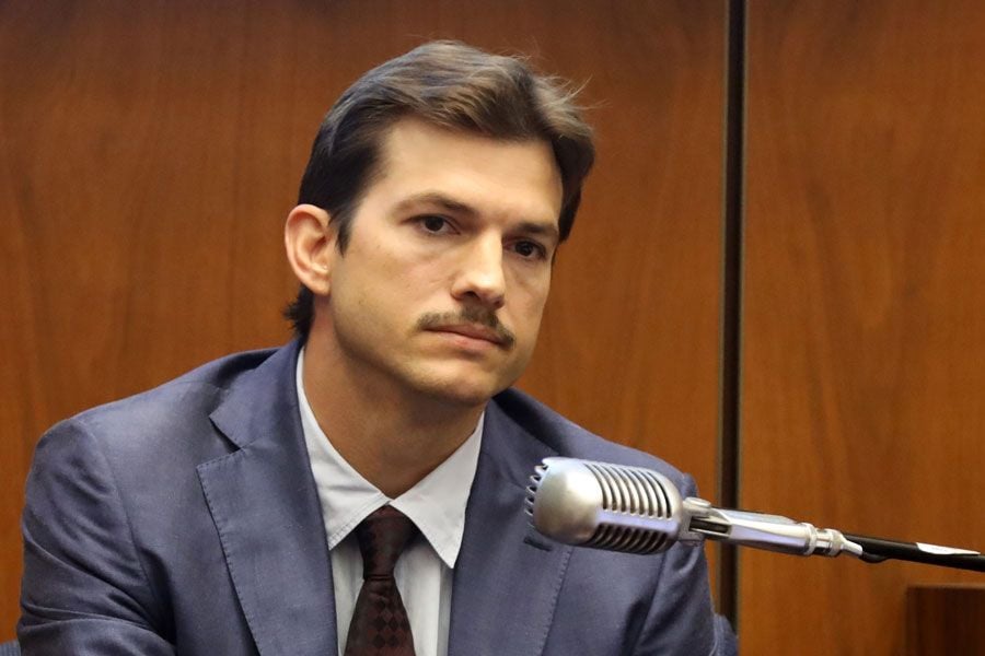 A man in a suit and tie with a mustache sits in a courtroom, looking serious, with a large microphone in front of him and wood-paneled walls in the background.