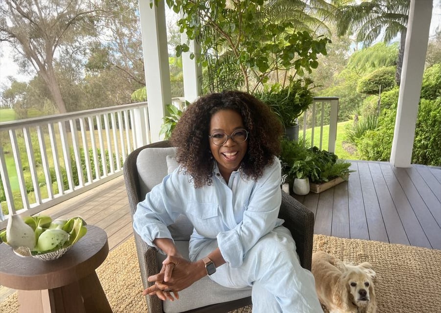 A woman with curly hair and glasses sits smiling on a porch chair, wearing a light blue outfit. A small dog lies beside her on a woven rug. The porch is surrounded by lush greenery and potted plants.