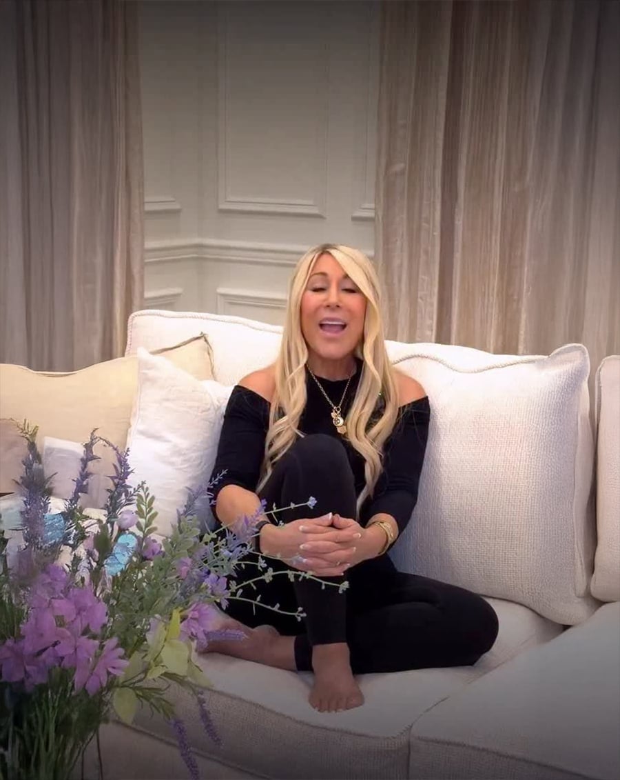 A woman with long blonde hair, wearing a black off-shoulder top and black pants, sits barefoot on a white couch with her legs crossed. Light purple flowers are in the foreground, and beige curtains hang behind her.