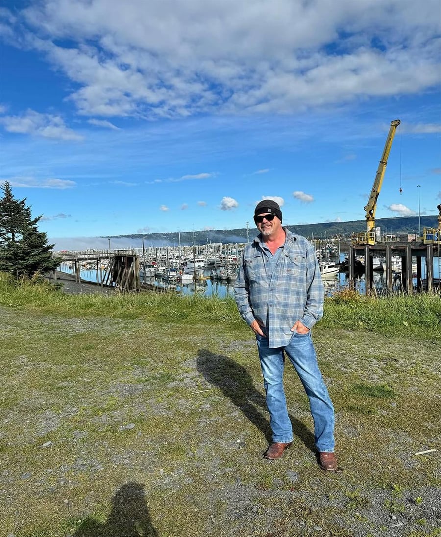 A man in a plaid shirt, jeans, and beanie stands on grass near a marina with boats, wooden docks, and a yellow crane. The sky is blue with scattered clouds.