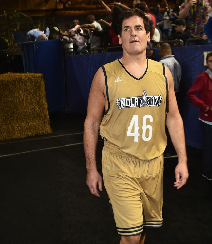 A man wearing a gold basketball uniform with the number 46 and "NOLA 17" on the front walks indoors, with people and hay bales visible in the background.