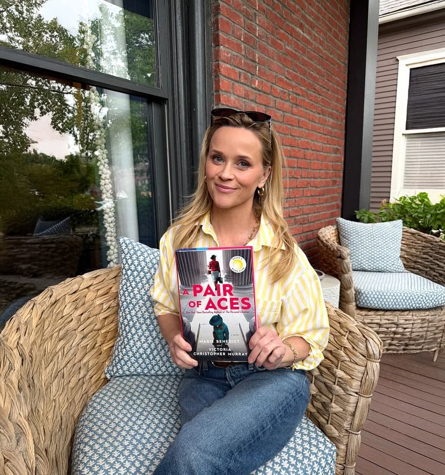 A smiling woman with blonde hair, wearing a yellow striped shirt and blue jeans, sits on a wicker chair on a porch holding a book titled "A Pair of Aces." Blue-patterned pillows are on the chair and neighboring couch.