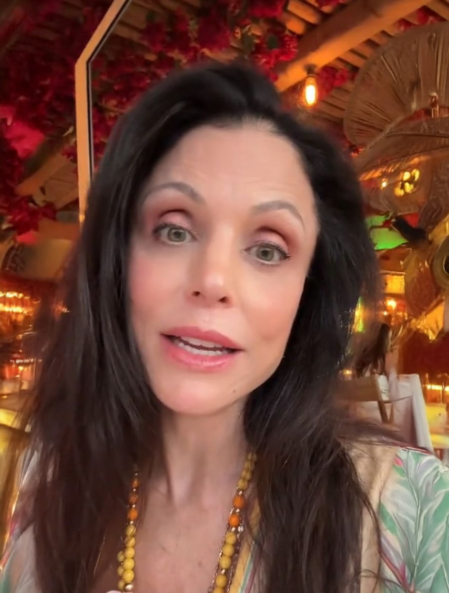 A woman with long dark hair and light eyes is indoors, wearing a beaded necklace and light makeup. Warm lighting and decorative elements, including red flowers and golden decor, are visible in the background.