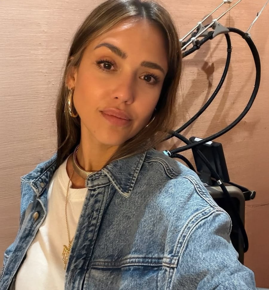 A woman with long brown hair wears a denim jacket over a white shirt, gold necklaces, and hoop earrings. She poses indoors in front of a beige wall with black cables and instruments visible behind her.