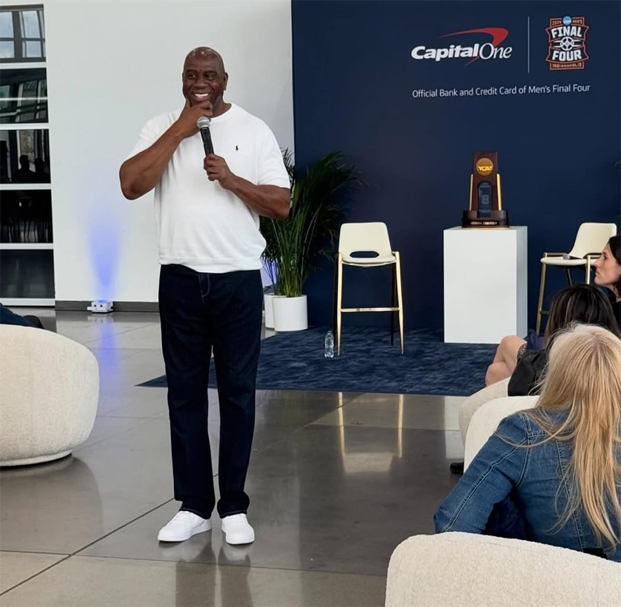 A man in a white shirt and dark pants speaks into a microphone, smiling, in front of a Capital One and NCAA Final Four backdrop, with seated audience members listening.