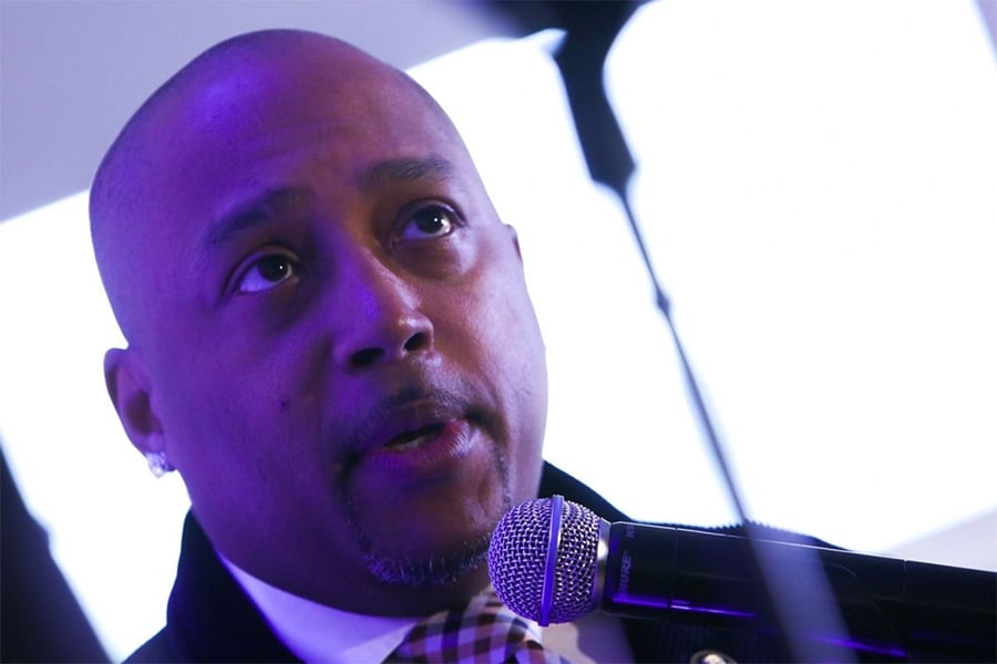 A man with a shaved head speaks into a microphone. He is wearing a dark jacket and a checkered shirt, and the background is brightly lit with purple tones on his face.