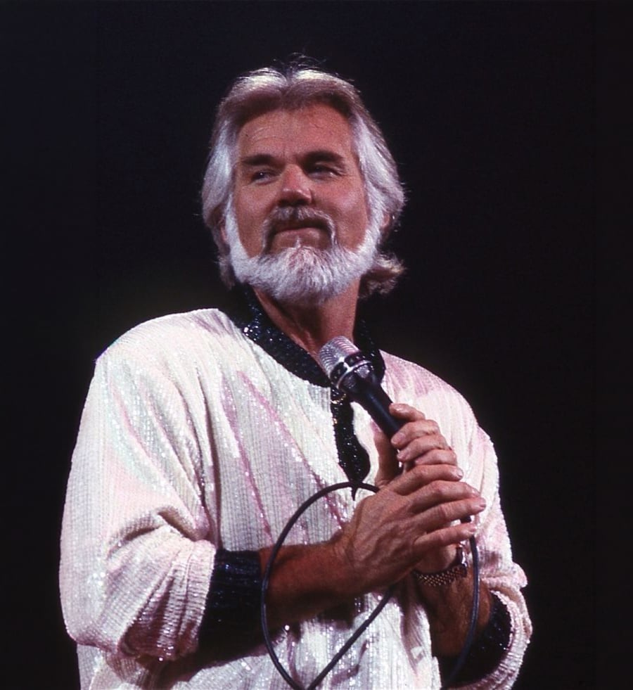A man with white hair and a beard stands on stage holding a microphone, wearing a sparkly light-colored shirt with a dark collar, against a dark background.