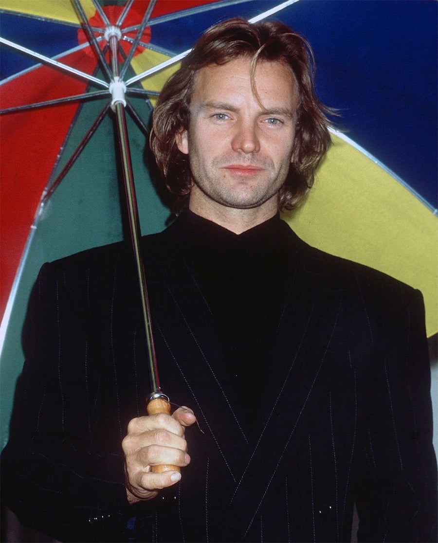 A man with shoulder-length hair, wearing a dark pinstripe suit, holds a multicolored umbrella and looks at the camera with a neutral expression.