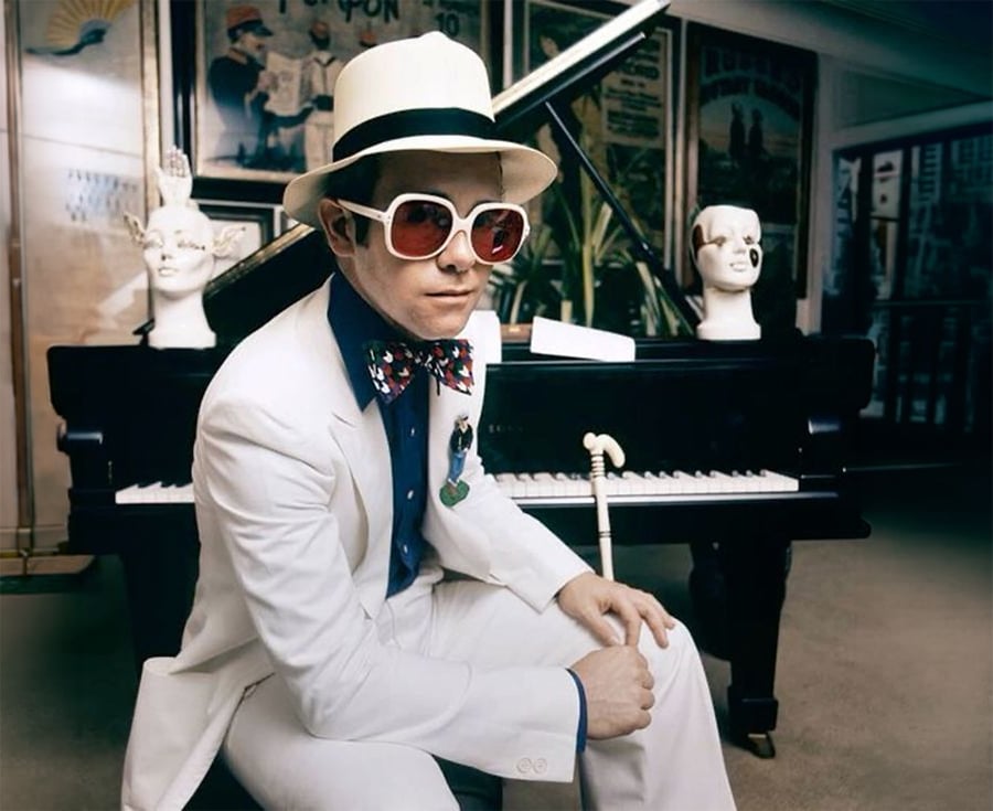 A person in a white suit, large sunglasses, and a white hat sits in front of a black piano. The piano has sculpted heads on it, and posters are visible in the background.