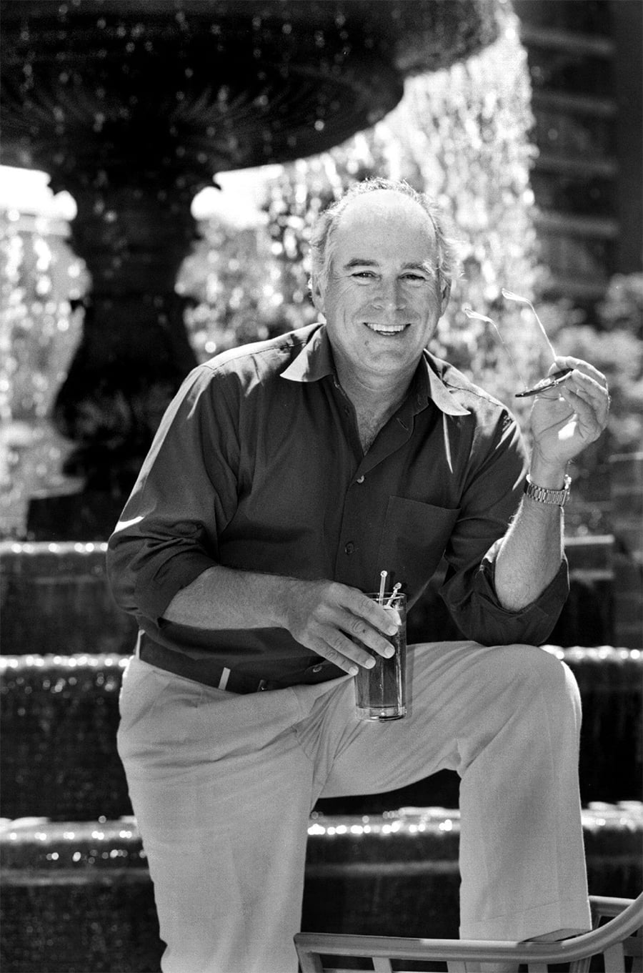 A smiling man in a button-up shirt and light pants stands in front of a fountain, holding a drink in one hand and sunglasses in the other, enjoying a sunny day.