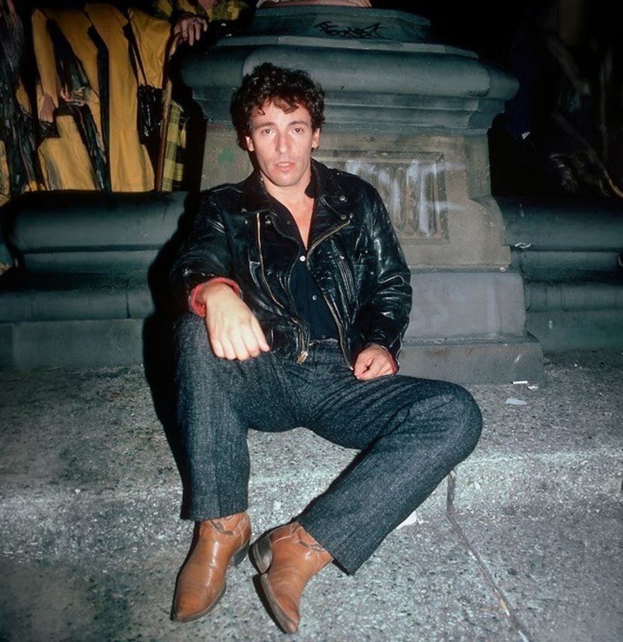 A man in a black leather jacket, dark pants, and brown boots sits on a concrete surface at night, leaning against a stone structure. People in yellow coats stand in the background.
