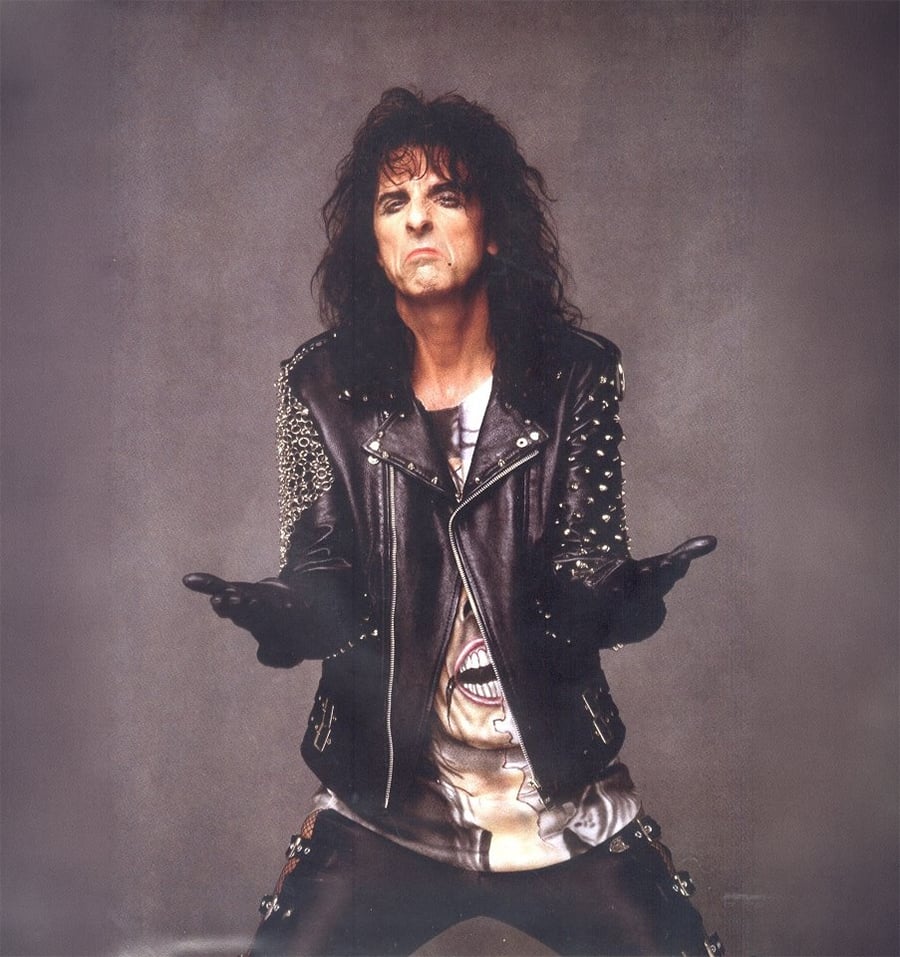 A person with long, dark, messy hair wears a black leather jacket with metal studs, black gloves, and dark pants, standing with hands outstretched and a serious expression against a plain background.