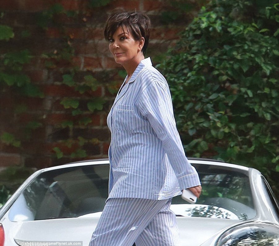 A woman with short dark hair, wearing striped blue and white pajamas, walks outdoors in front of a silver convertible car, holding a phone, with green foliage and a brick wall in the background.