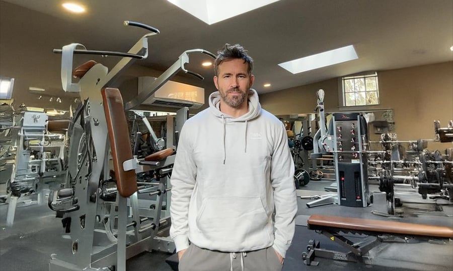 A man with short brown hair and a beard, wearing a light gray hoodie and dark pants, stands in the middle of a spacious gym filled with various exercise machines and weights. Natural light comes in through skylights above.