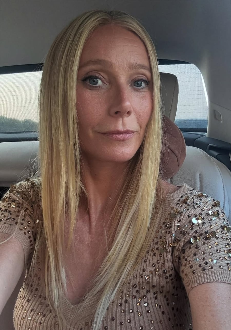 A woman with long blonde hair and blue eyes sits in the back seat of a car, wearing a beige top decorated with sequins. Sunlight illuminates her face as she looks into the camera, giving a slight smile.