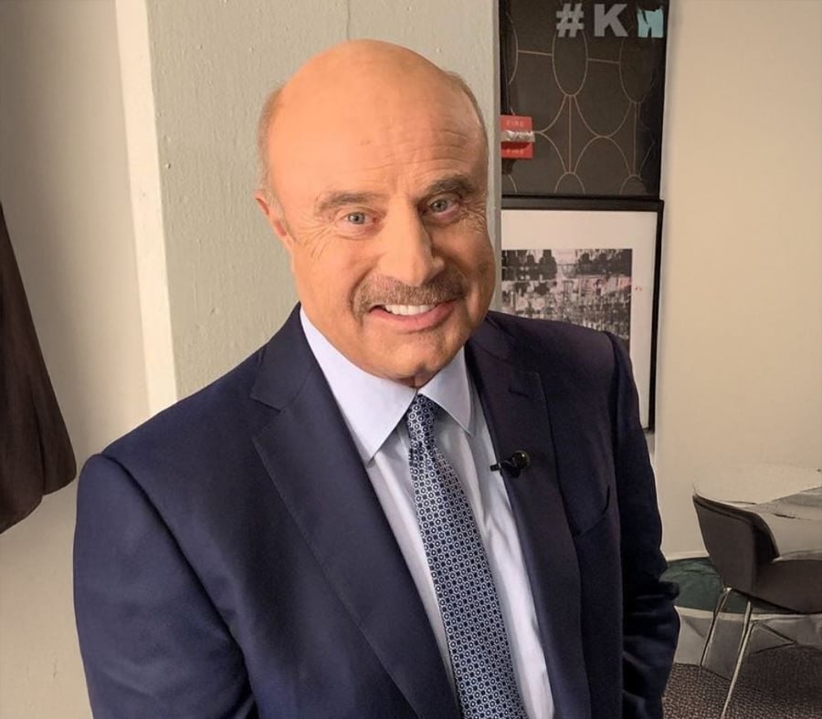 A bald man with a mustache is smiling at the camera, wearing a navy suit, light blue shirt, and patterned tie, standing indoors near a table and framed photos on the wall.