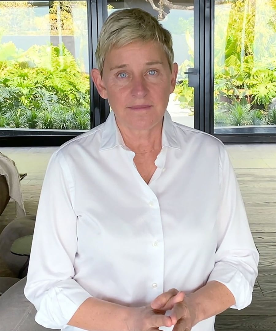 A person with short light hair wearing a white button-up shirt stands indoors, hands clasped, with large windows and greenery visible in the background.