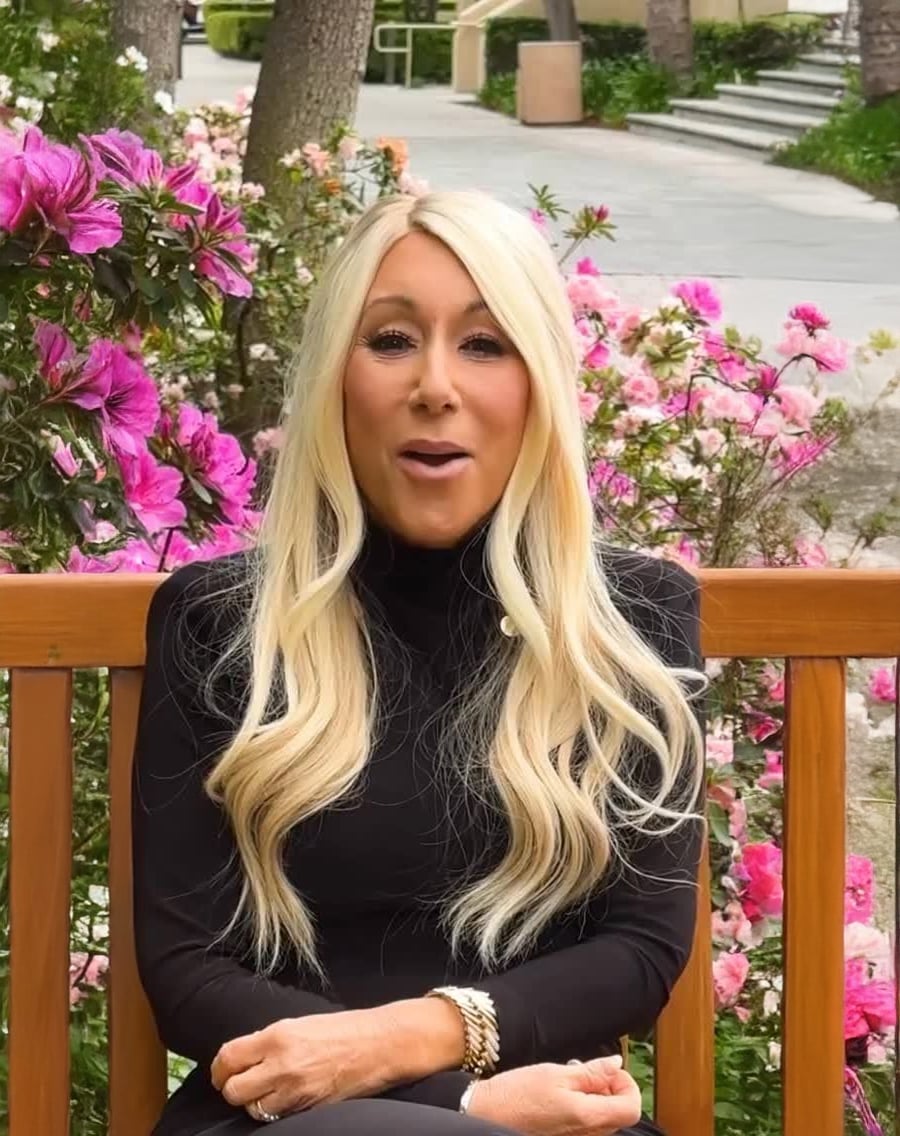 A woman with long blonde hair, wearing a black turtleneck dress, sits on a wooden bench surrounded by vibrant pink flowers and greenery. She is smiling, with her arms crossed in her lap.
