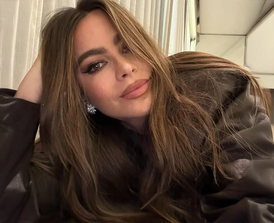 A woman with long brown hair and light makeup rests her head on her hand, wearing a dark jacket and sparkling earrings, posing indoors near light-colored curtains.