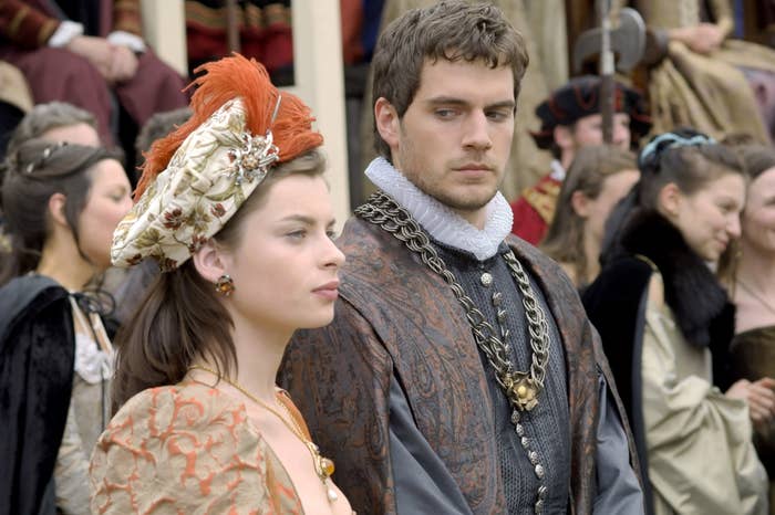 Two actors in period costumes with ornate patterns, one wearing a feathered hat, appear in a historical film scene