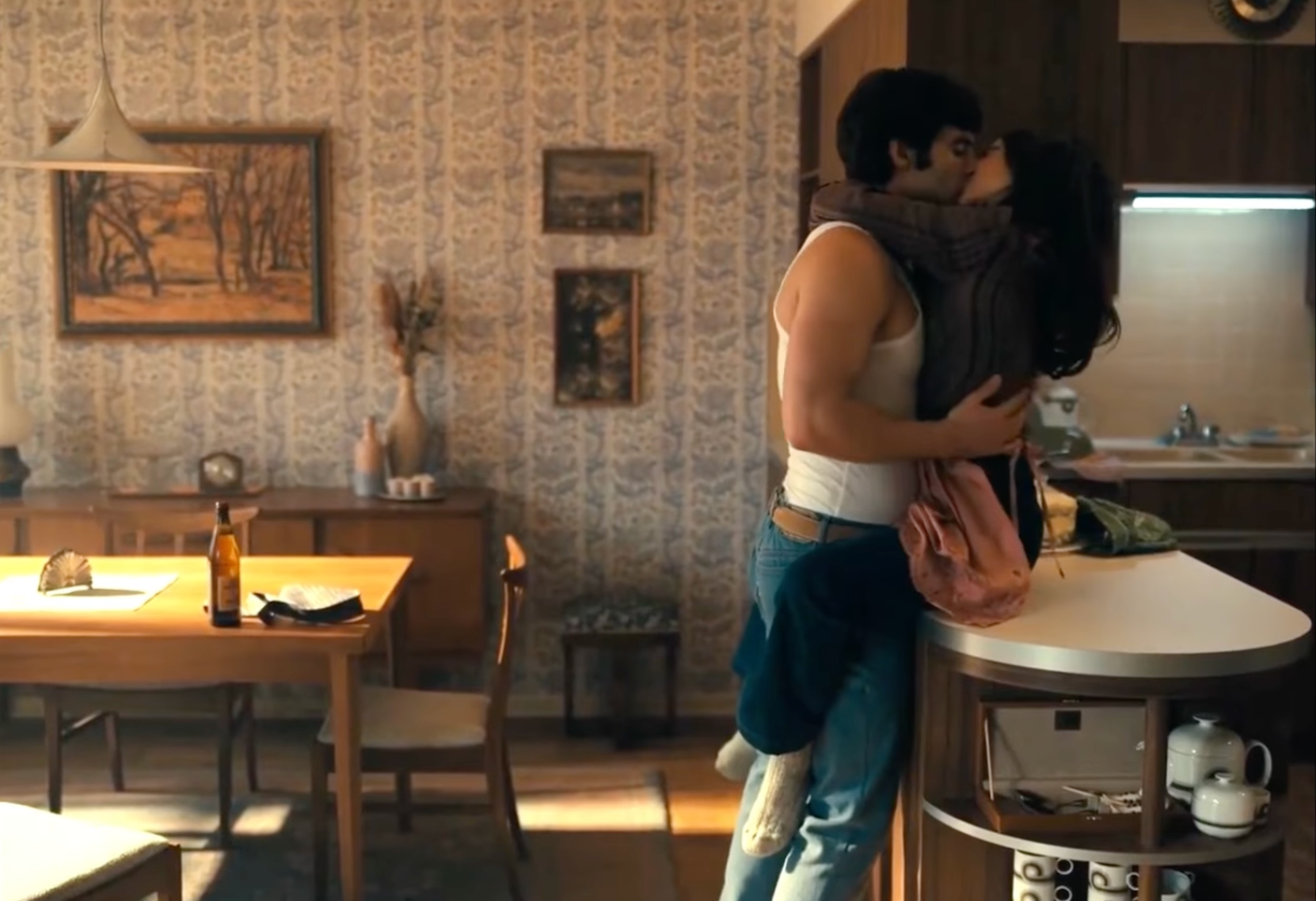 A man and Emilia Clark share a passionate kiss in a cozy, vintage-style kitchen