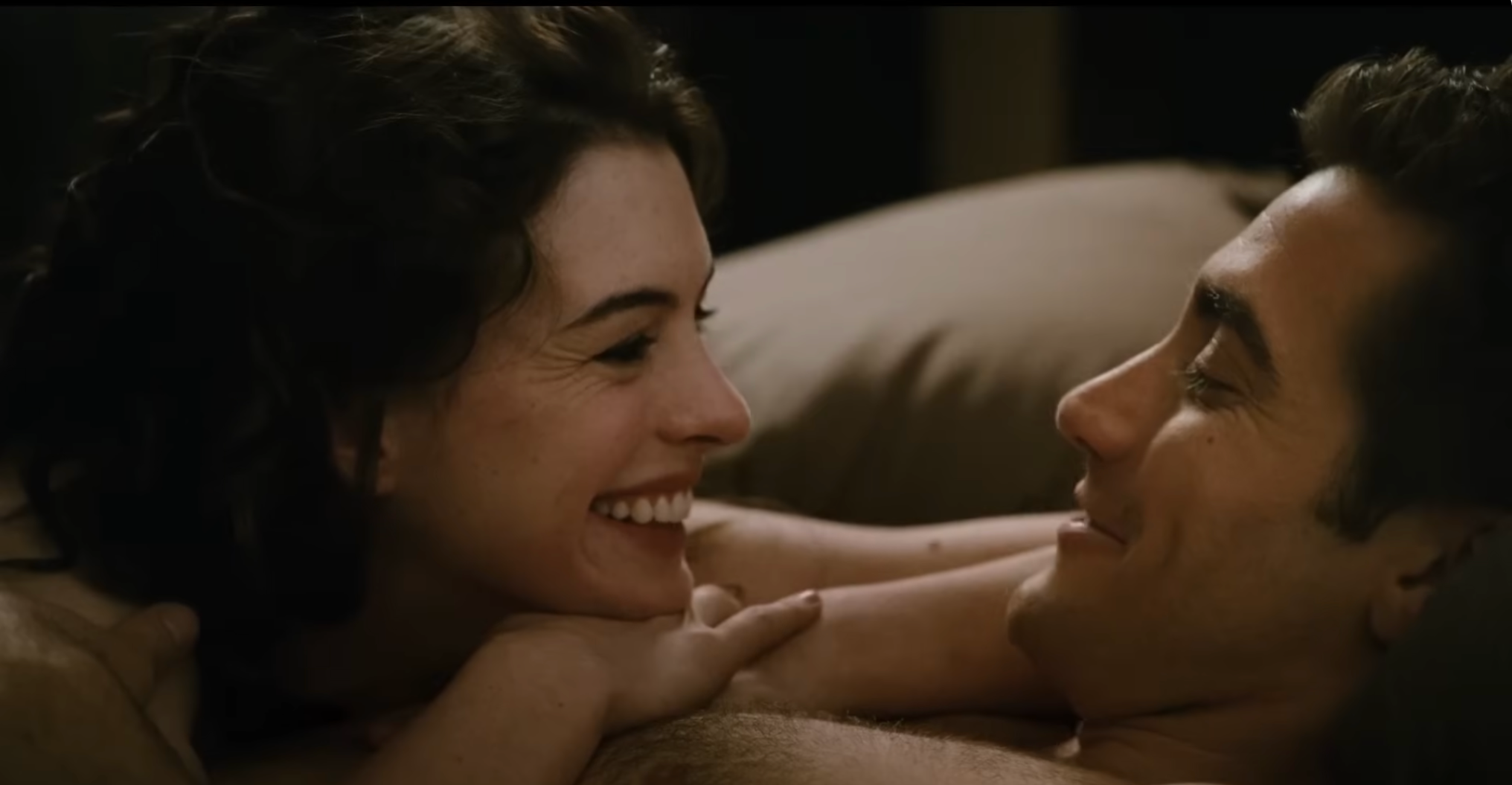 Anne Hathaway and Jake Gyllenhaal are lying closely together in bed, smiling at each other