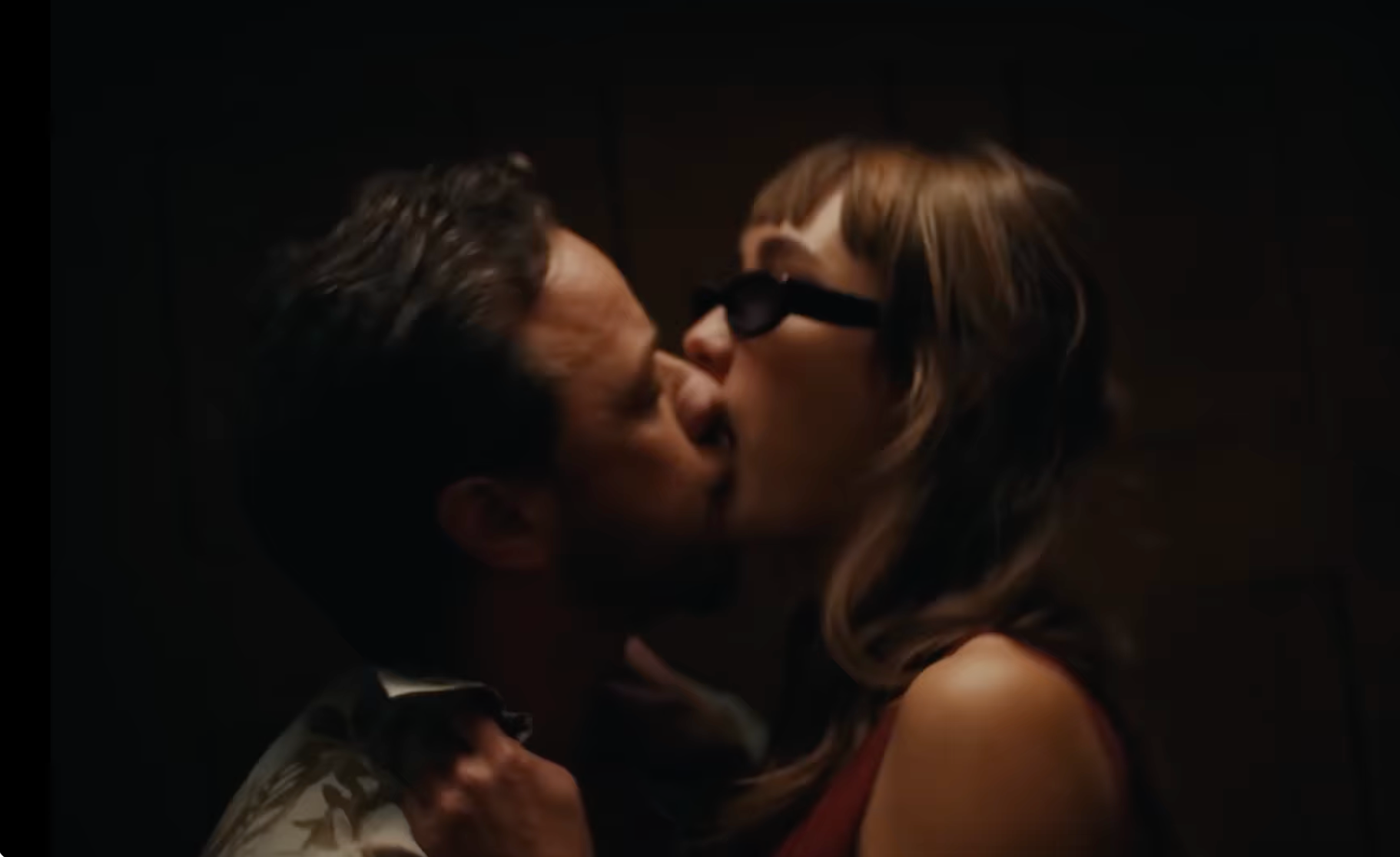 A couple shares a passionate kiss in a dimly lit setting. The woman is wearing sunglasses and has long hair