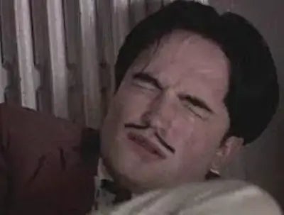 Robert Pattinson, with a mustache, squinting and wearing a suit, appears to react emotionally