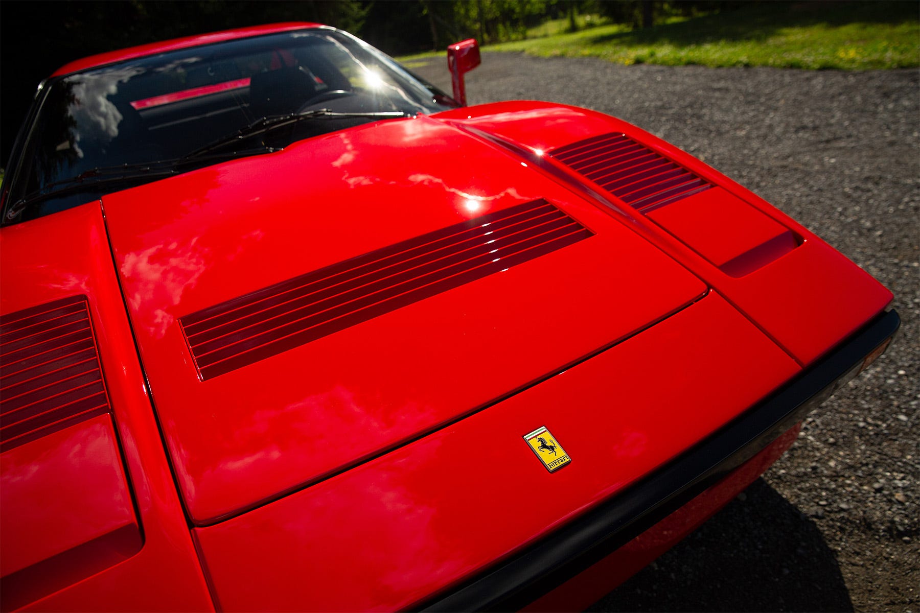 hauba crvenog sportskog automobila s ferrarijevim logom, postavljena naspram šljunčanog prilaza i drveća.