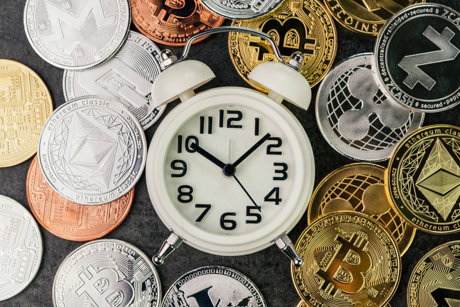 An overhead shot of a white analog alarm clock with black numbers and hands, set to approximately 10:10. The clock is centered and surrounded by a diverse array of metallic cryptocurrency coins in gold, silver, and copper hues, resting on a dark, textured surface.