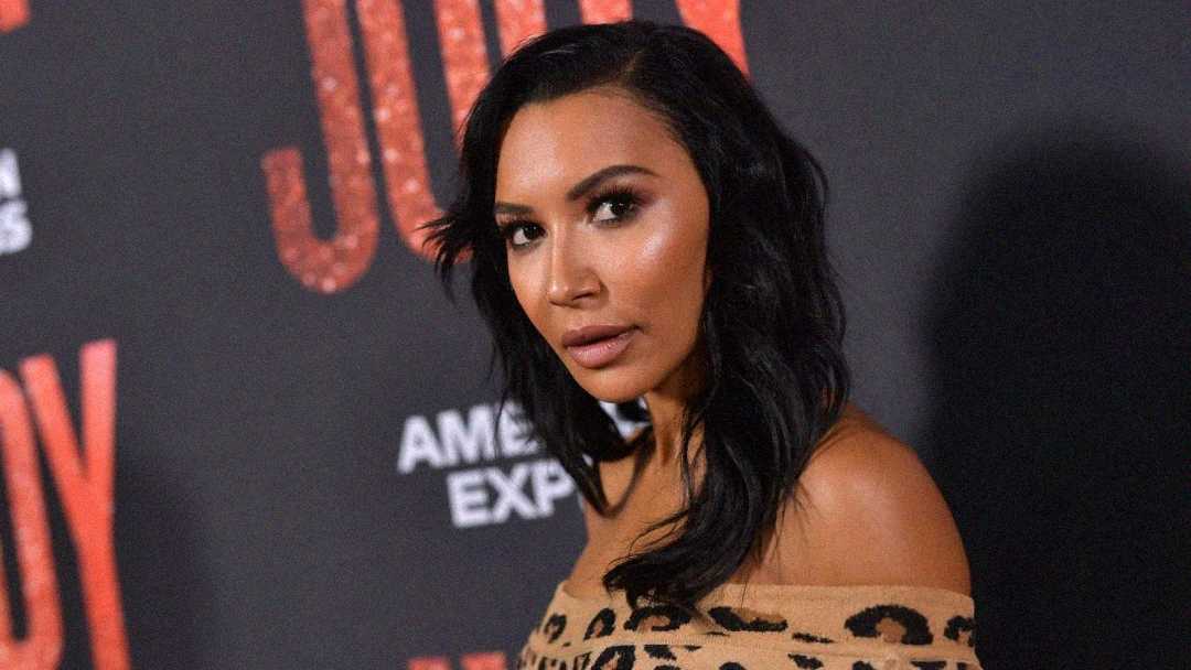 Una mujer con cabello oscuro, largo y ondulado, que lleva una blusa con estampado de leopardo y hombros descubiertos, posa frente a un fondo con un texto rojo brillante y el logotipo de American Express.