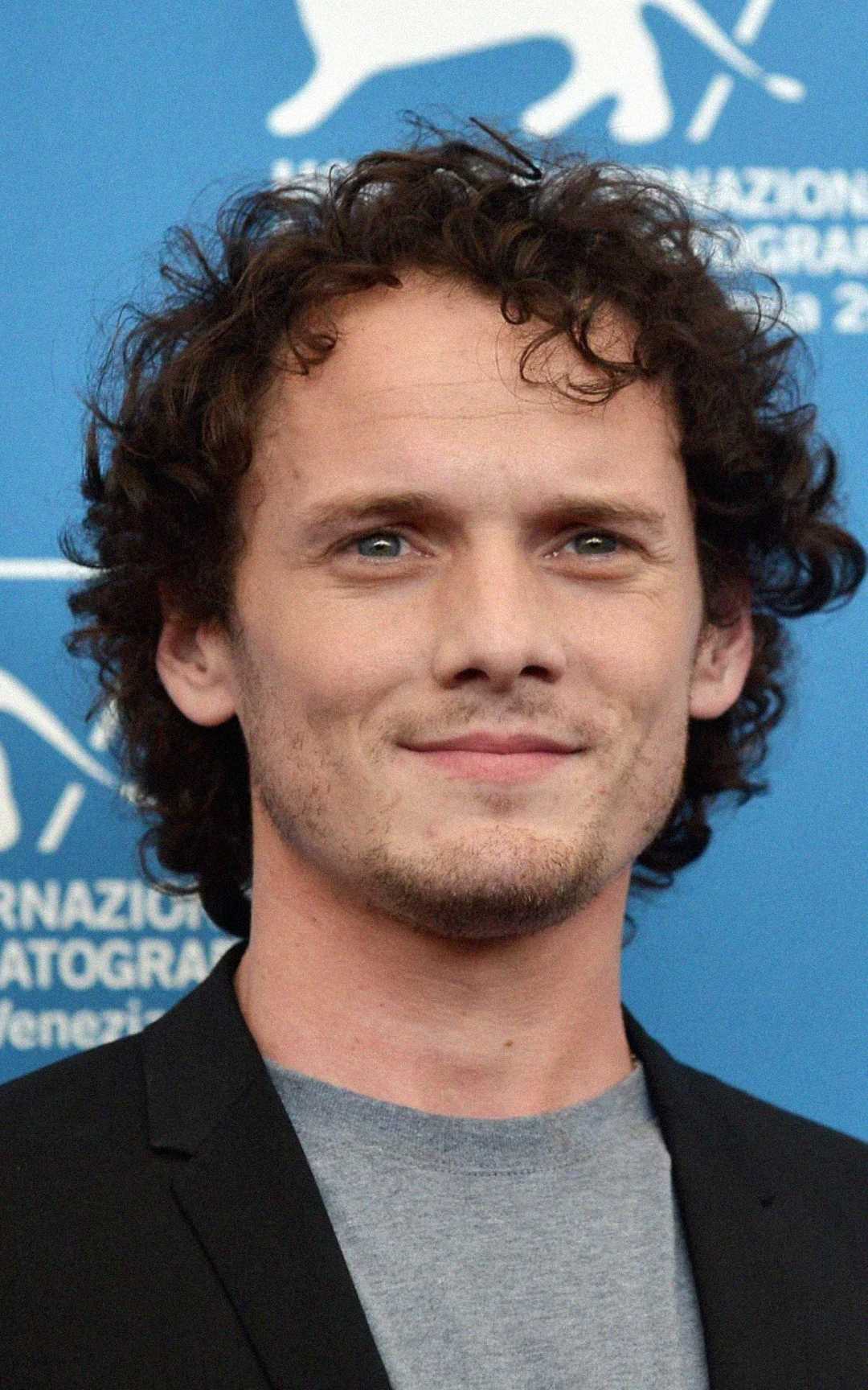 Un hombre de cabello castaño rizado y ojos azules sonríe levemente. Lleva una camisa gris y una chaqueta negra, de pie frente a un fondo azul con texto blanco y el logo de un león.